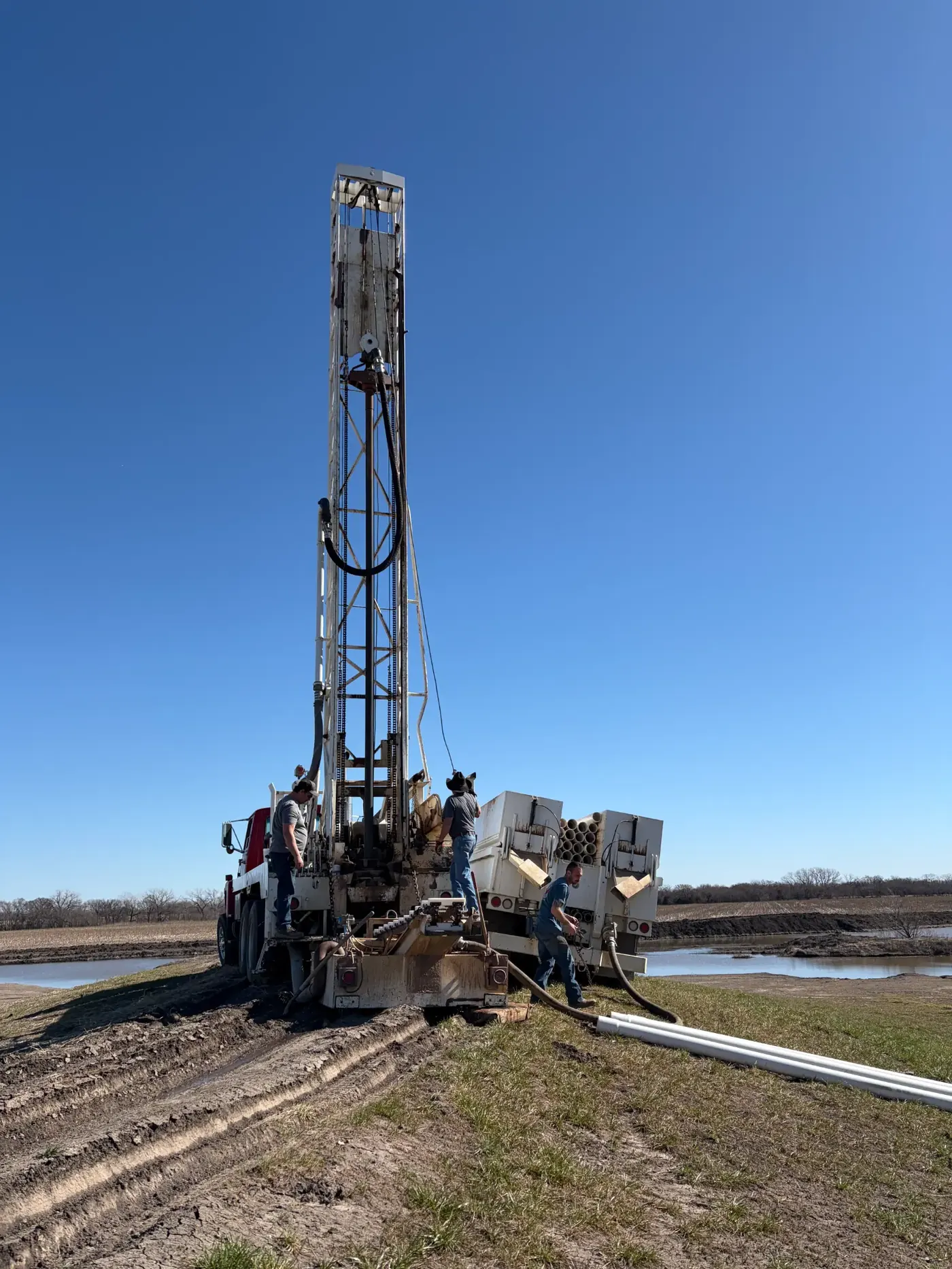 Chase Drilling LLC drilling rig operating in a flat Kansas agricultural field near a water feature, drilling a new irrigation well for crop or landscape watering